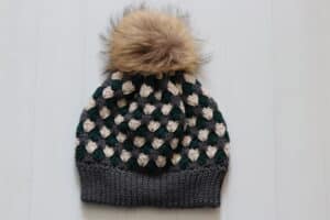 A Crochet Beanie worked in the nesting shell stitch worked in green, grey and white yarn with a tan coloured faux fur pompom