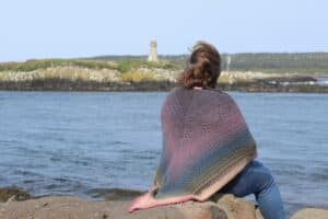 Woman sitting on rocky shore wearing the Brier Island Crochet Shawl