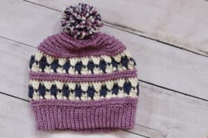 The Charlottetown Beanie worked in shades of purple yarn and white featuring a hand made pompom