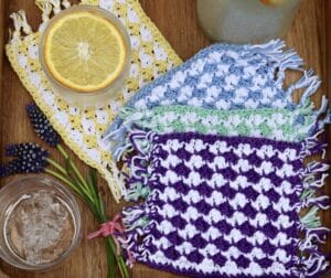 A set of four crochet coasters displayed on a summer themed table with lemonade
